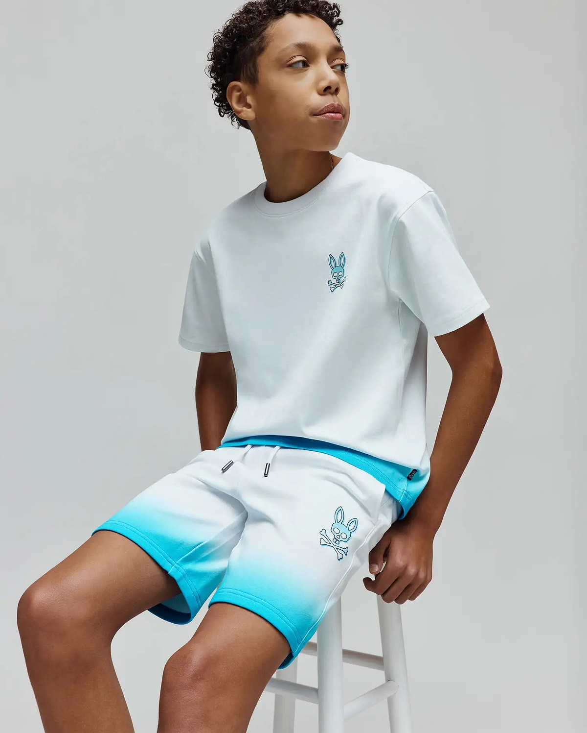 White jersey t-shirt with small blue embroidered Psycho Bunny skull logo on chest, paired with KIDS DAX SWEATSHORT featuring white to blue ombre, relaxed fit, and large blue embroidered bunny skull on thigh.