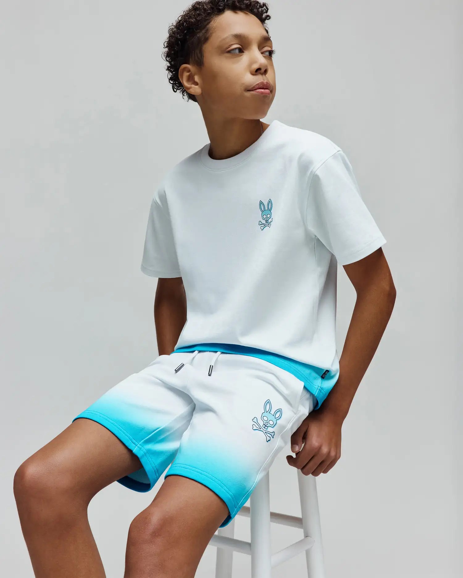 White jersey t-shirt with small blue embroidered Psycho Bunny skull logo on chest, paired with KIDS DAX SWEATSHORT featuring white to blue ombre, relaxed fit, and large blue embroidered bunny skull on thigh.