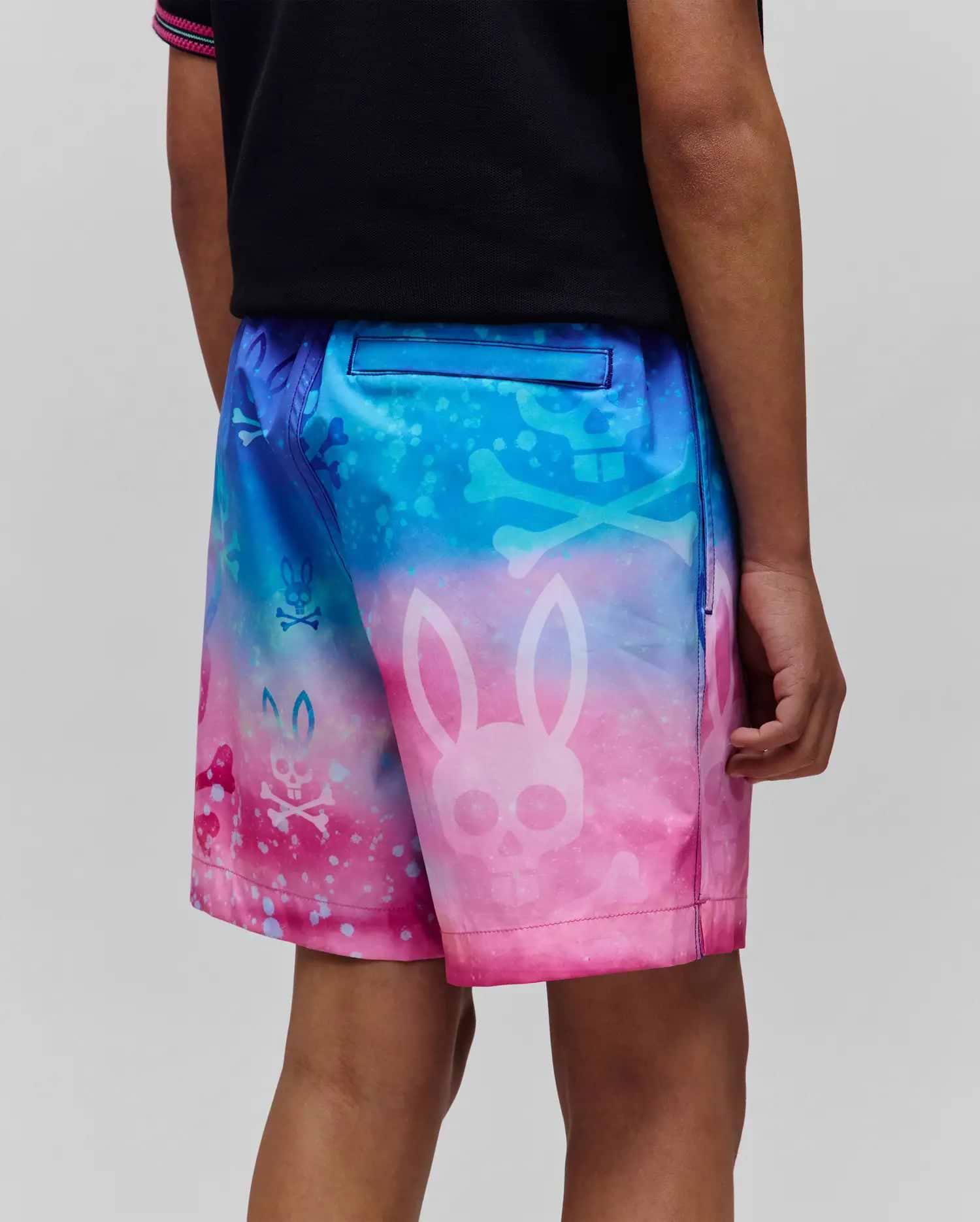 Kids Caspian Swim Trunk in a vibrant blue to pink gradient, featuring an all-over print of large and small Psycho Bunny skull and crossbones logos with a splatter effect.