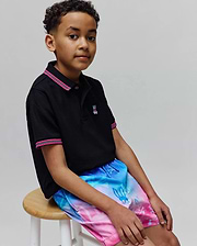 Black pique polo with pink/white striped collar and cuffs, featuring a small embroidered Psycho Bunny logo, paired with Kids Caspian Swim Trunk in a blue-to-pink gradient with allover bunny skull pattern.