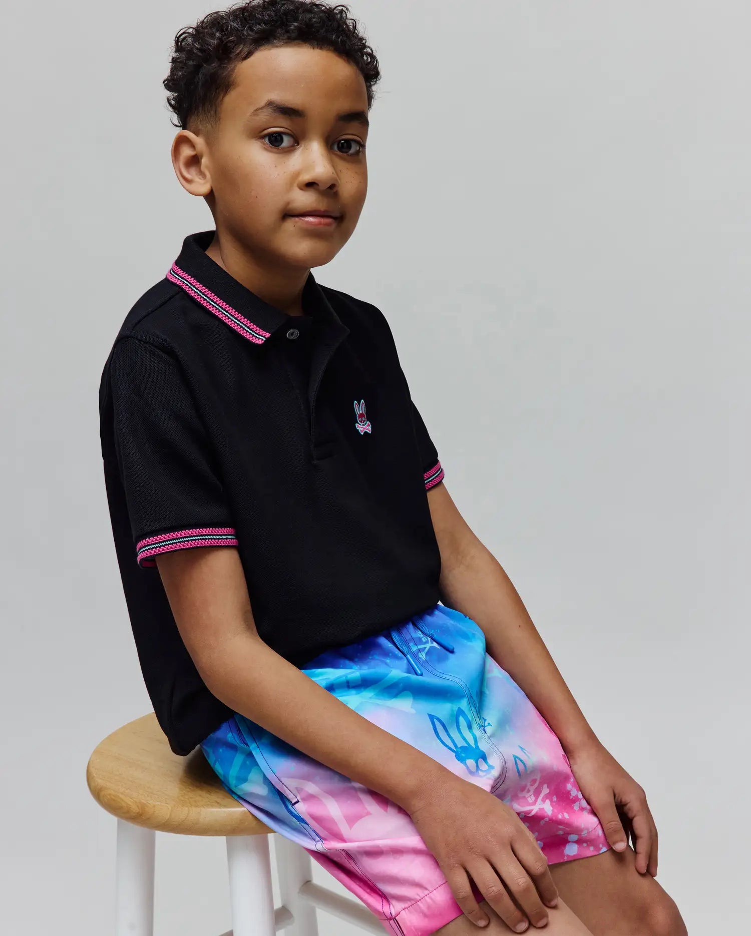 Black pique polo with pink/white striped collar and cuffs, featuring a small embroidered Psycho Bunny logo, paired with Kids Caspian Swim Trunk in a blue-to-pink gradient with allover bunny skull pattern.