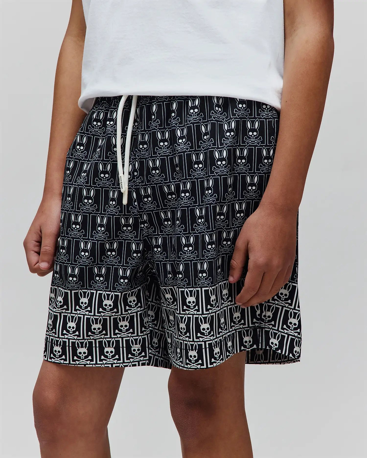 Kids Keon Swim Trunk, black relaxed-fit swim shorts featuring an all-over white Psycho Bunny skull and crossbones pattern, a distinct border design, and a white drawstring with Psycho Bunny text.