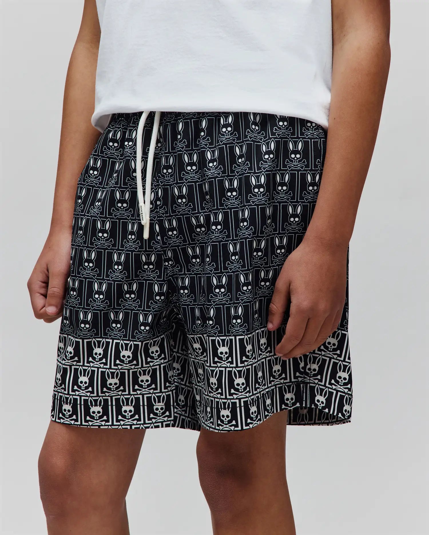 Kids Keon Swim Trunk, black relaxed-fit swim shorts featuring an all-over white Psycho Bunny skull and crossbones pattern, a distinct border design, and a white drawstring with Psycho Bunny text.