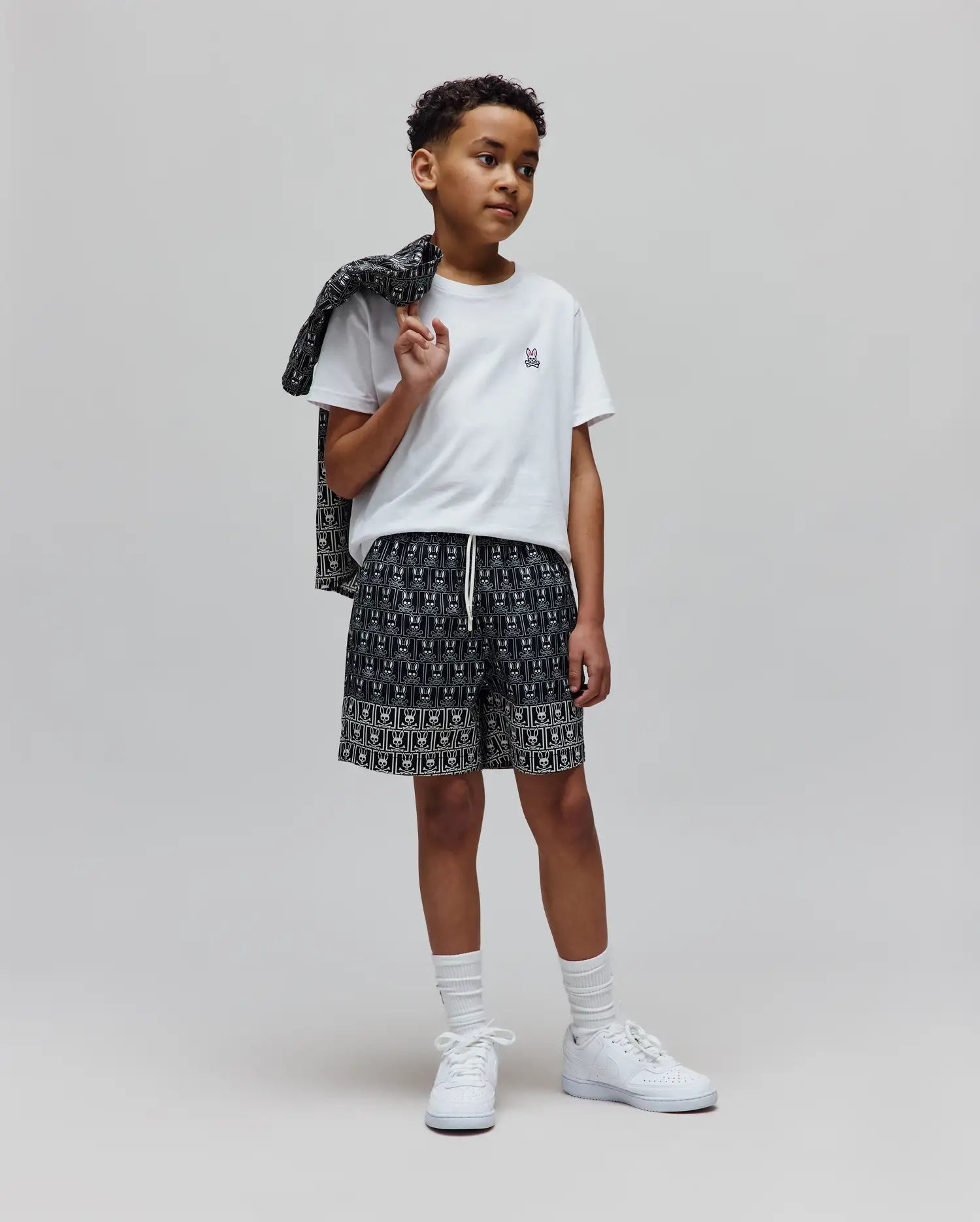 Classic fit white jersey t-shirt with small embroidered Psycho Bunny logo, Kids Keon Swim Trunk in black with all-over white skull pattern, and matching patterned jacket.