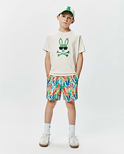 Off-white jersey t-shirt with a large green Psycho Bunny skull graphic, paired with multicolor KIDS TANNER SWIM TRUNK featuring an all-over bunny skull print, and a cream baseball cap with an embroidered green bunny logo.