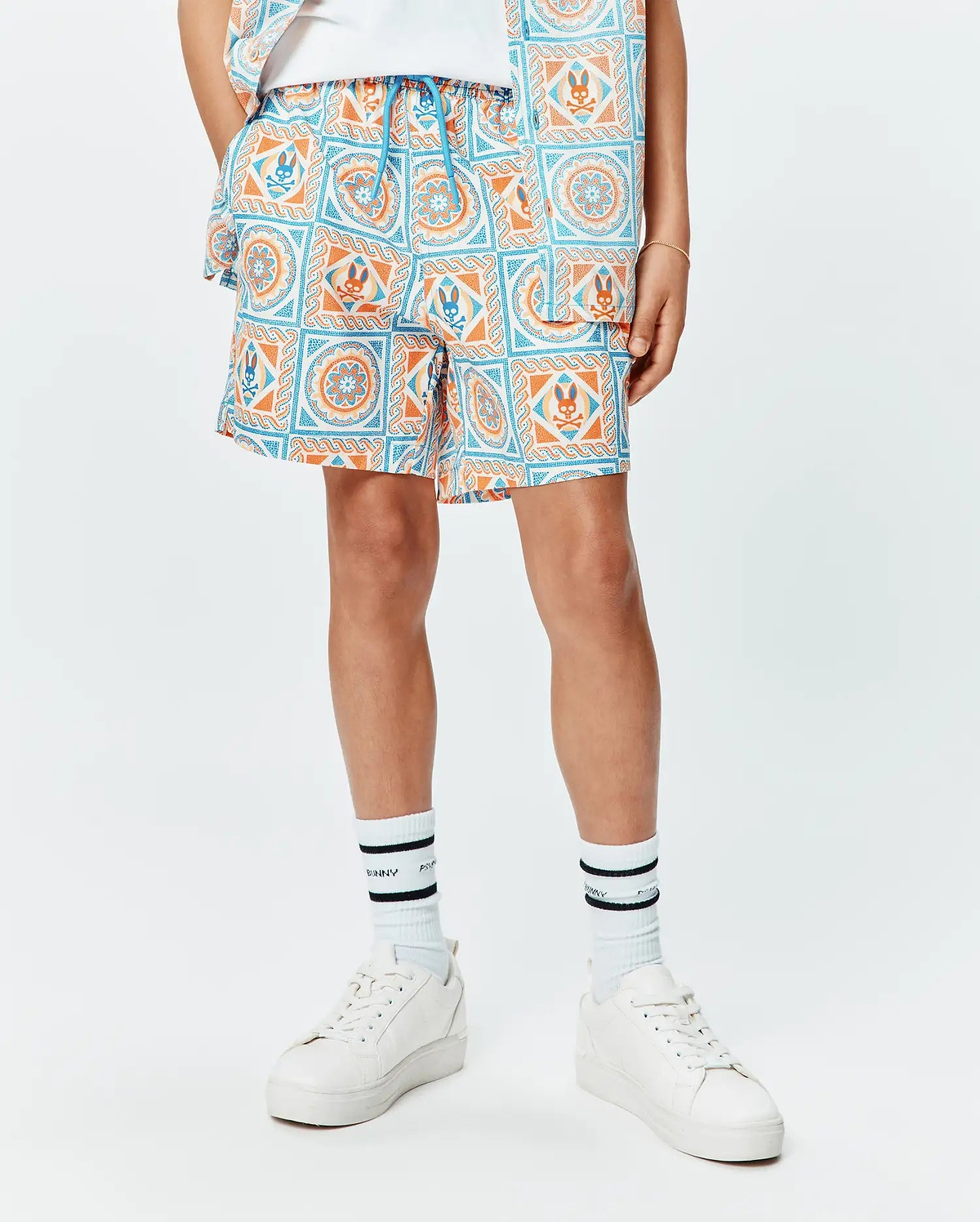 Kids Frank Swim Trunk, a relaxed fit swim trunk featuring a vibrant white, blue, and orange tiled pattern with repeated Psycho Bunny skull logos and a blue drawstring.