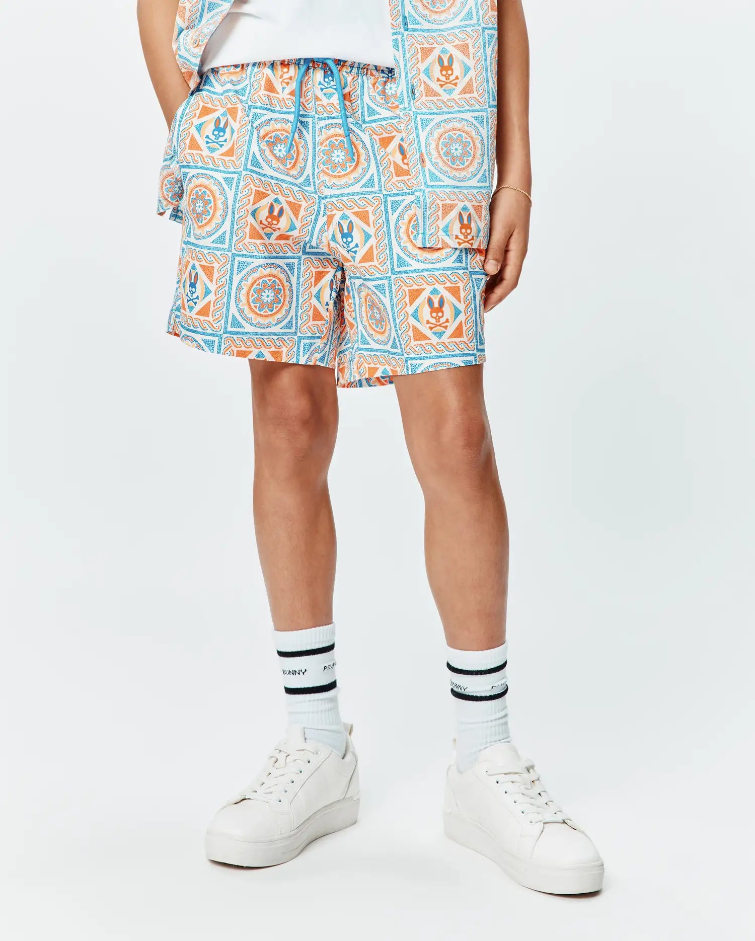 Kids Frank Swim Trunk, a relaxed fit swim trunk featuring a vibrant white, blue, and orange tiled pattern with repeated Psycho Bunny skull logos and a blue drawstring.