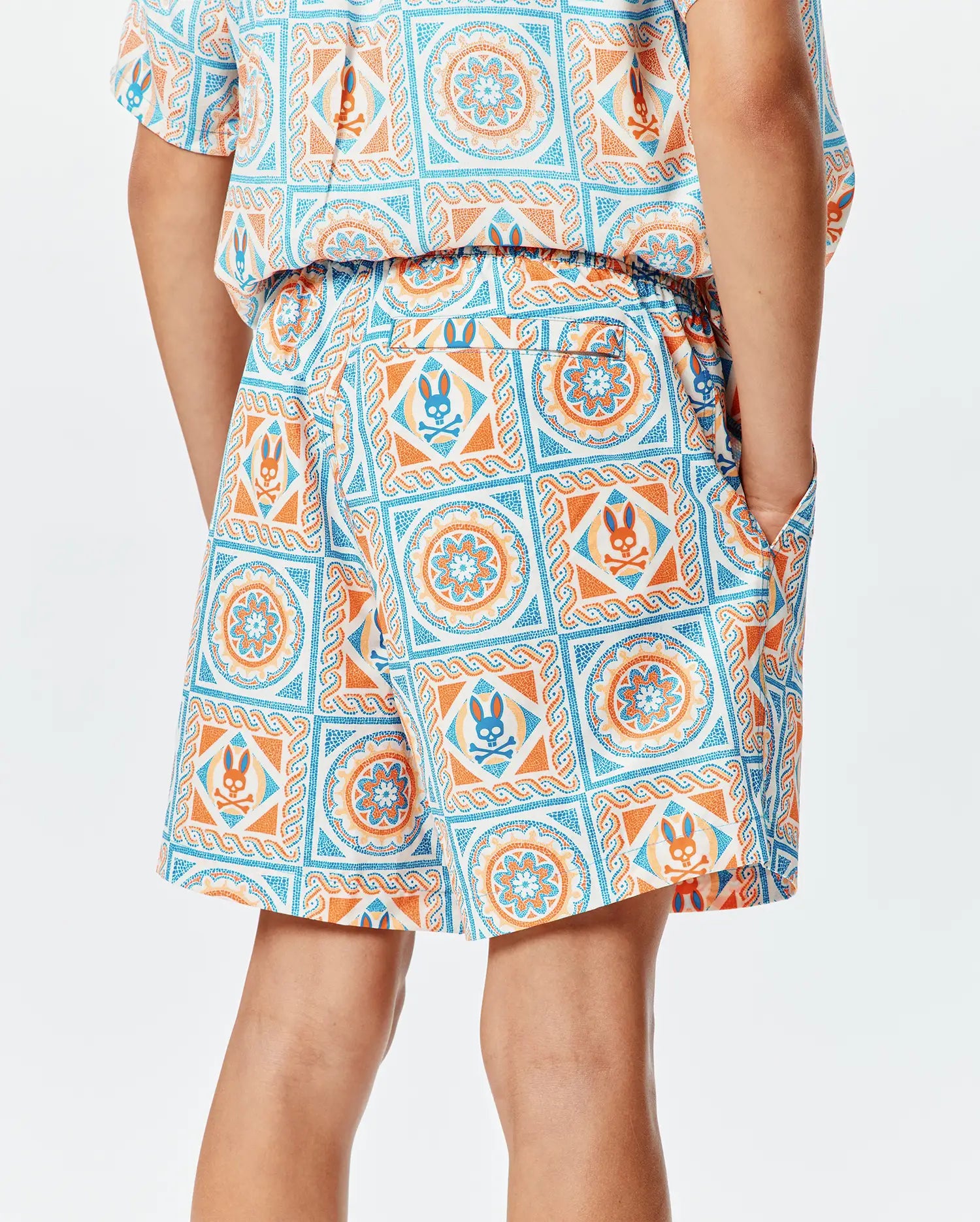 Kids Frank Swim Trunk in a relaxed fit, featuring a vibrant white, orange, and blue mosaic tile pattern with integrated Psycho Bunny skull logos, elastic waistband, and side pockets.