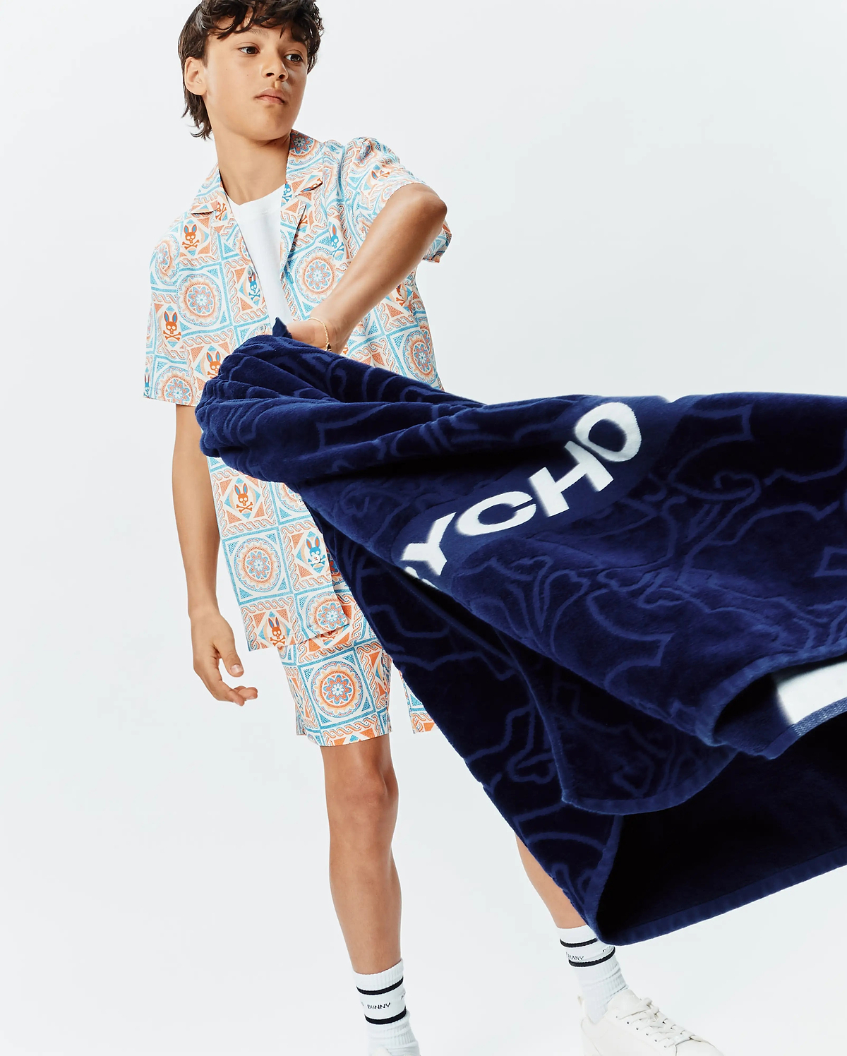Kids Frank Swim Trunk and matching camp shirt in a vibrant blue and orange mosaic pattern featuring Psycho Bunny skull logos. Relaxed fit. Paired with a navy terry towel displaying large white PSYCHO text.