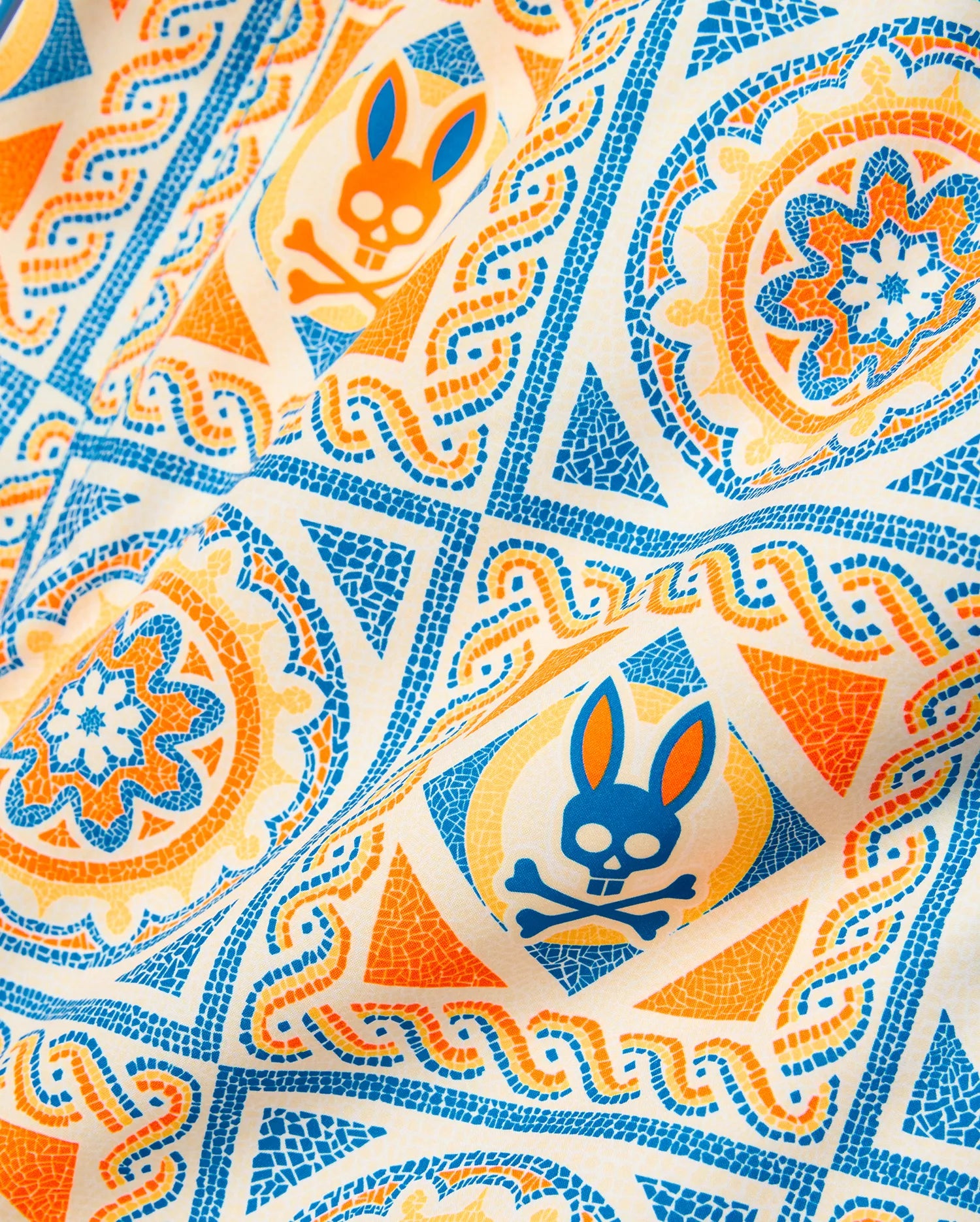 Kids Frank Swim Trunk in white with an all-over mosaic pattern featuring blue, orange, and yellow geometric shapes, floral motifs, and the iconic Psycho Bunny skull logo.