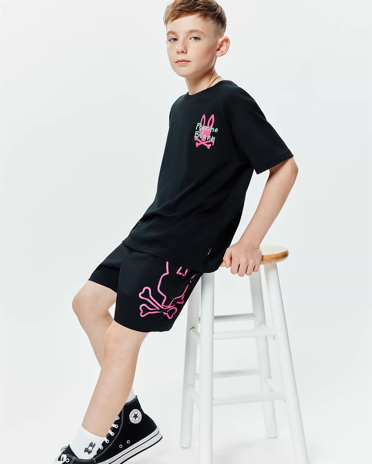 Black jersey t-shirt with a relaxed fit, featuring a neon pink and green Psycho Bunny logo on the chest. Paired with black Kids Duncan Bunny Print Swim Trunk, showcasing a large neon pink bunny skull and crossbones graphic on the left leg.
