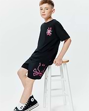 Black jersey t-shirt with a relaxed fit, featuring a neon pink and green Psycho Bunny logo on the chest. Paired with black Kids Duncan Bunny Print Swim Trunk, showcasing a large neon pink bunny skull and crossbones graphic on the left leg.