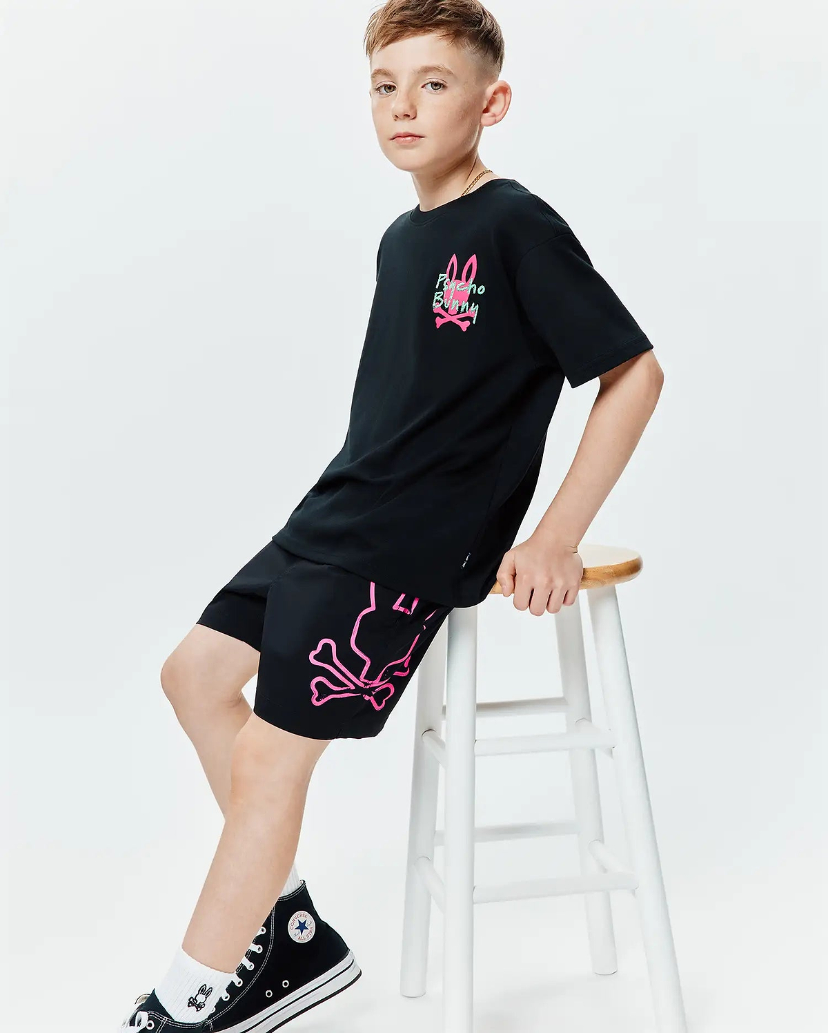 Black jersey t-shirt with a relaxed fit, featuring a neon pink and green Psycho Bunny logo on the chest. Paired with black Kids Duncan Bunny Print Swim Trunk, showcasing a large neon pink bunny skull and crossbones graphic on the left leg.