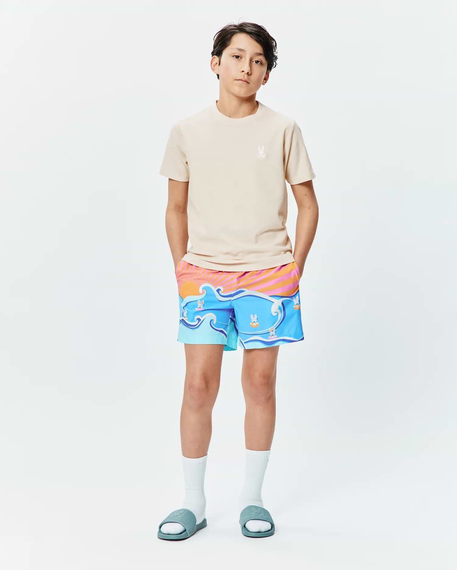 Beige jersey t-shirt with small embroidered Psycho Bunny logo on left chest, paired with Kids Ray Swim Trunk 6 featuring a vibrant blue wave and sunset print with floating bunny skull logos.