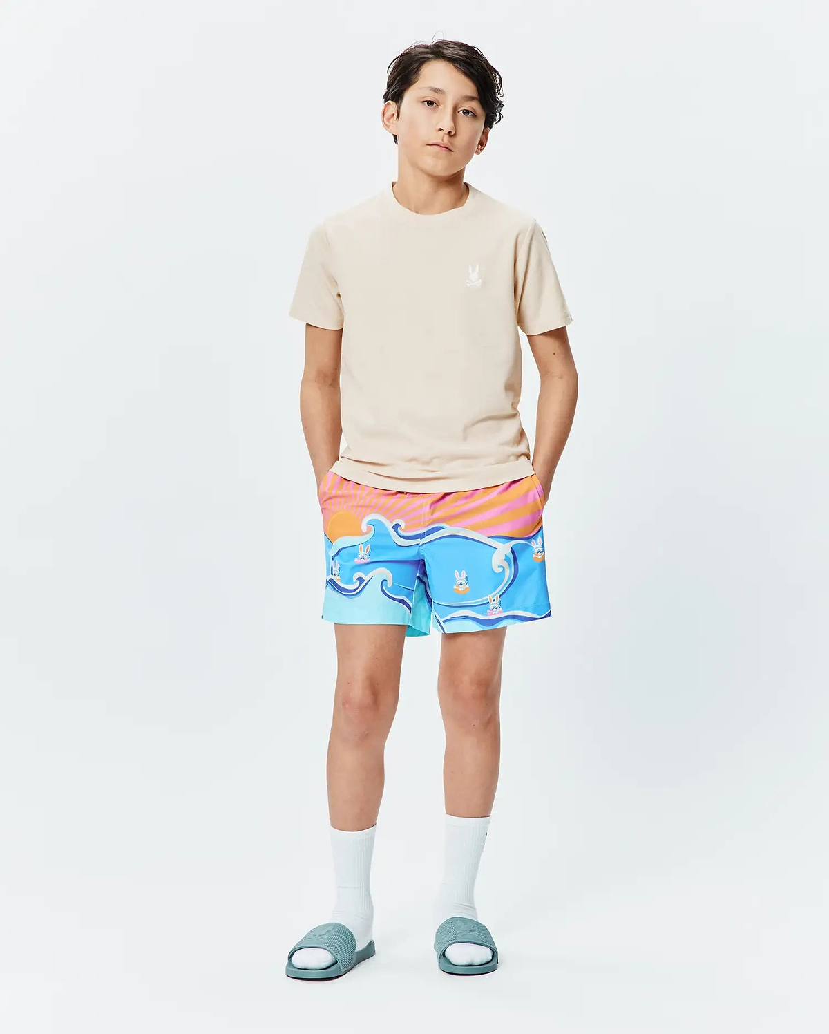 Beige jersey t-shirt with small embroidered Psycho Bunny logo on left chest, paired with Kids Ray Swim Trunk 6 featuring a vibrant blue wave and sunset print with floating bunny skull logos.