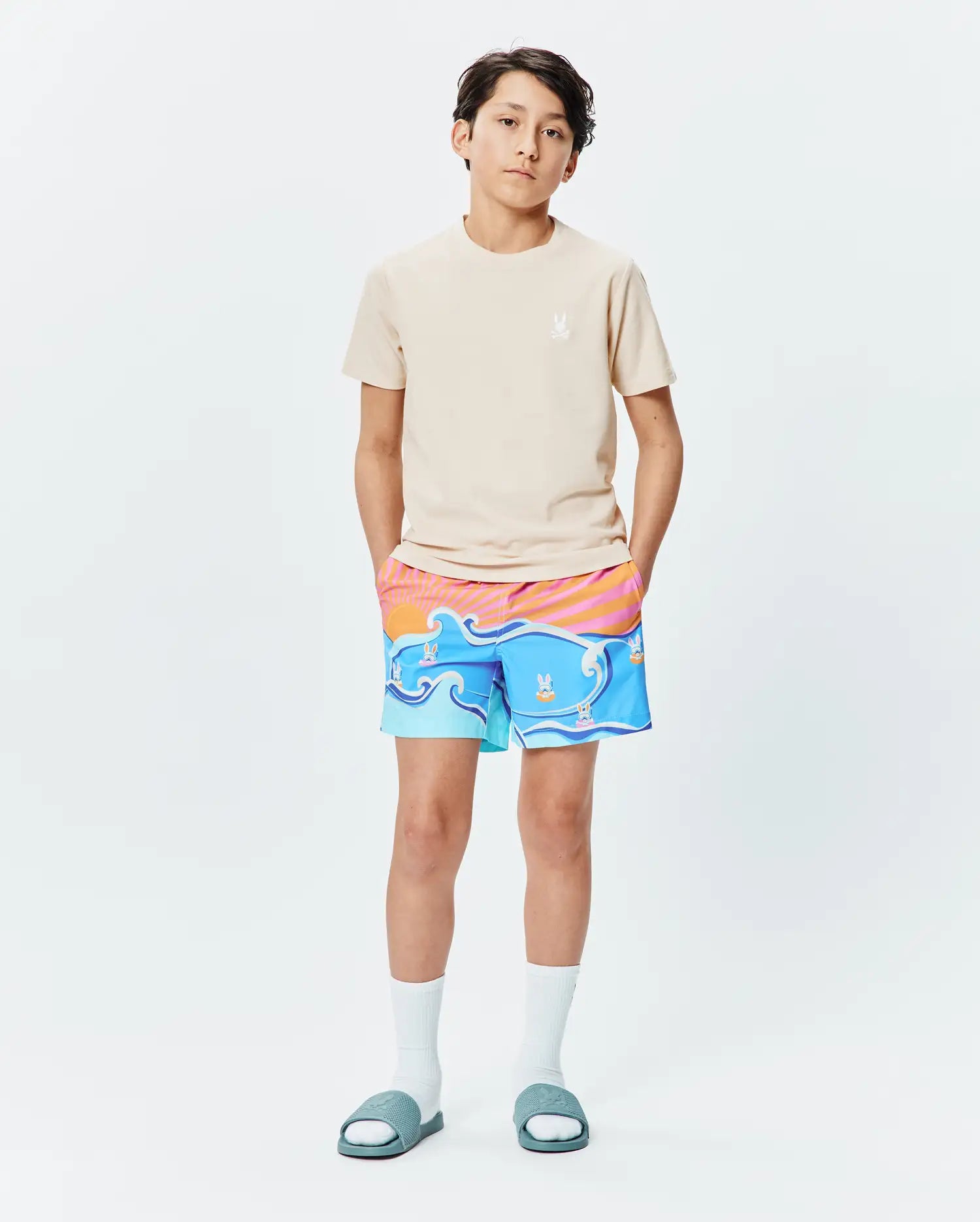 Beige jersey t-shirt with small embroidered Psycho Bunny logo on left chest, paired with Kids Ray Swim Trunk 6 featuring a vibrant blue wave and sunset print with floating bunny skull logos.