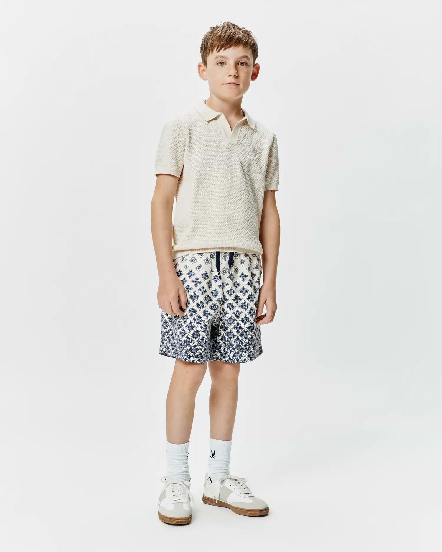 Classic fit cream pique knit polo with embroidered Psycho Bunny logo on chest, paired with Kids Tobias Swim Trunk featuring a white base and intricate blue geometric pattern, drawstring waist.