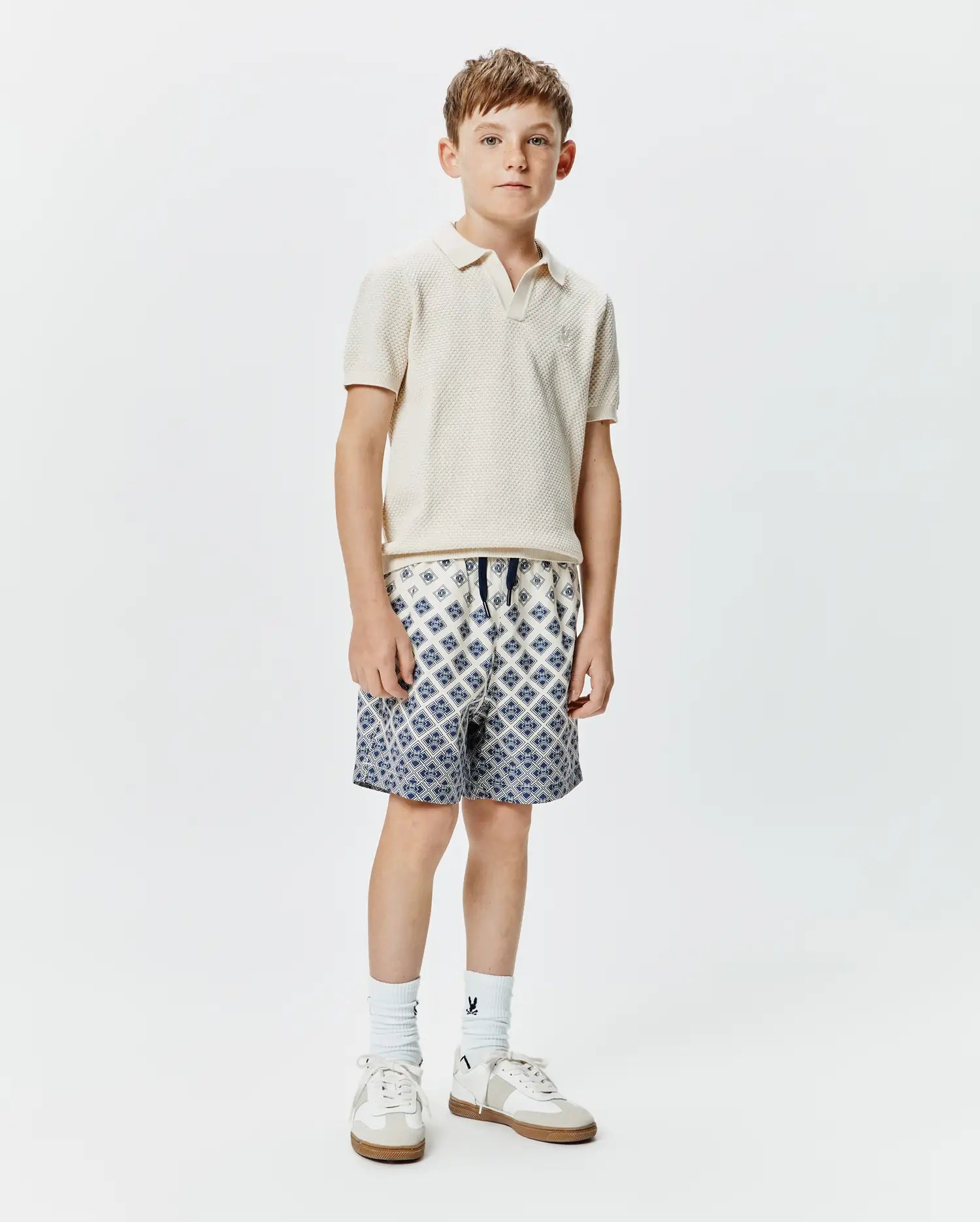 Classic fit cream pique knit polo with embroidered Psycho Bunny logo on chest, paired with Kids Tobias Swim Trunk featuring a white base and intricate blue geometric pattern, drawstring waist.