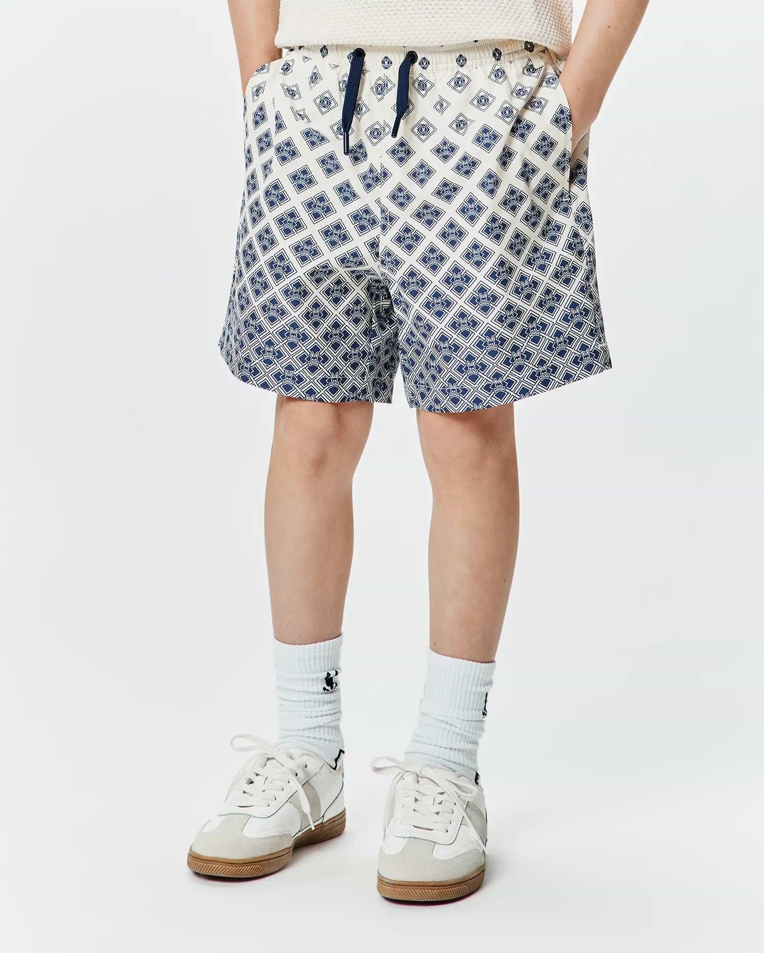 Kids Tobias Swim Trunk, cream with a navy blue all-over geometric pattern of the iconic Psycho Bunny skull logo, relaxed fit, smooth quick-dry fabric, and a navy drawstring.