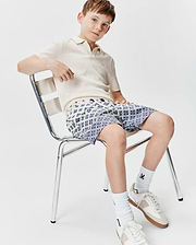 Cream textured knit polo with a small embroidered Psycho Bunny logo on the chest, paired with Kids Tobias Swim Trunk featuring an all-over blue and white geometric pattern with subtle bunny skulls.