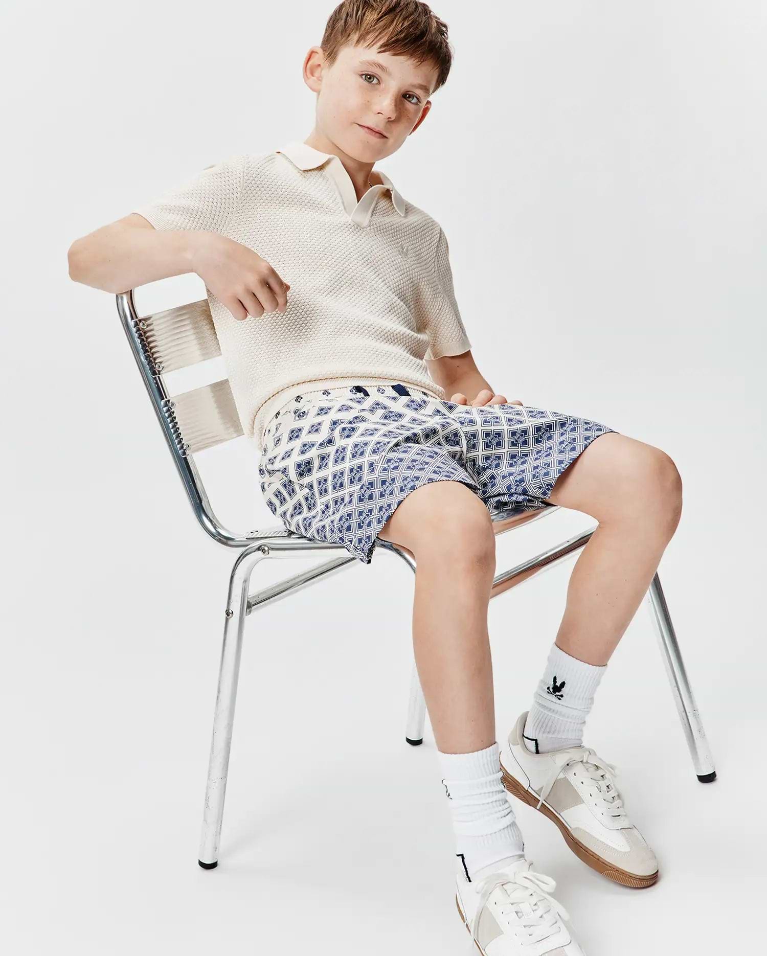 Cream textured knit polo with a small embroidered Psycho Bunny logo on the chest, paired with Kids Tobias Swim Trunk featuring an all-over blue and white geometric pattern with subtle bunny skulls.