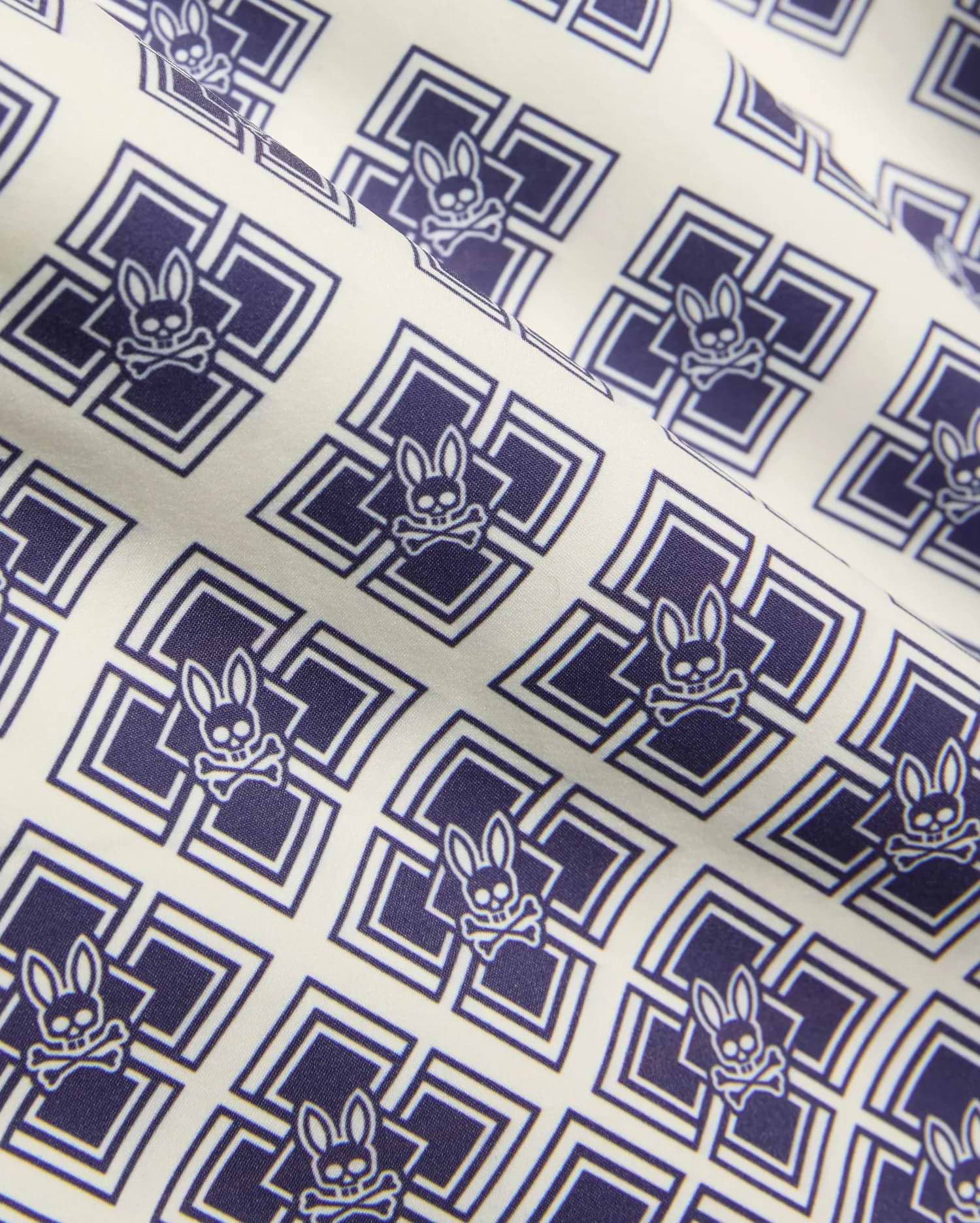Kids Tobias Swim Trunk in cream with an all-over navy blue geometric square pattern featuring repeated Psycho Bunny skull logos on smooth woven fabric.