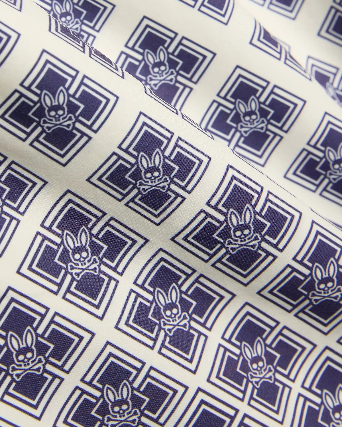 Kids Tobias Swim Trunk in cream with an all-over navy blue geometric square pattern featuring repeated Psycho Bunny skull logos on smooth woven fabric.