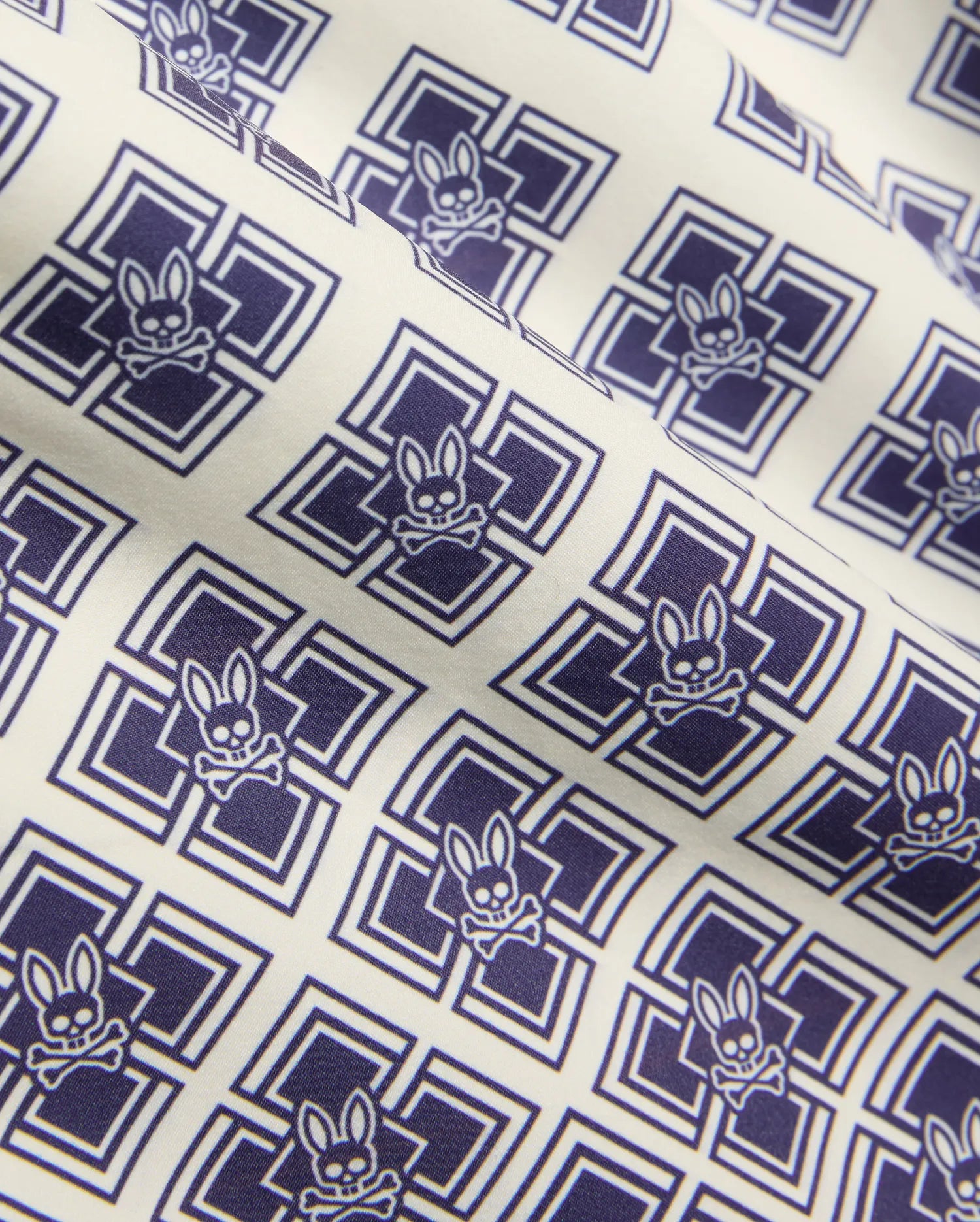 Kids Tobias Swim Trunk in cream with an all-over navy blue geometric square pattern featuring repeated Psycho Bunny skull logos on smooth woven fabric.