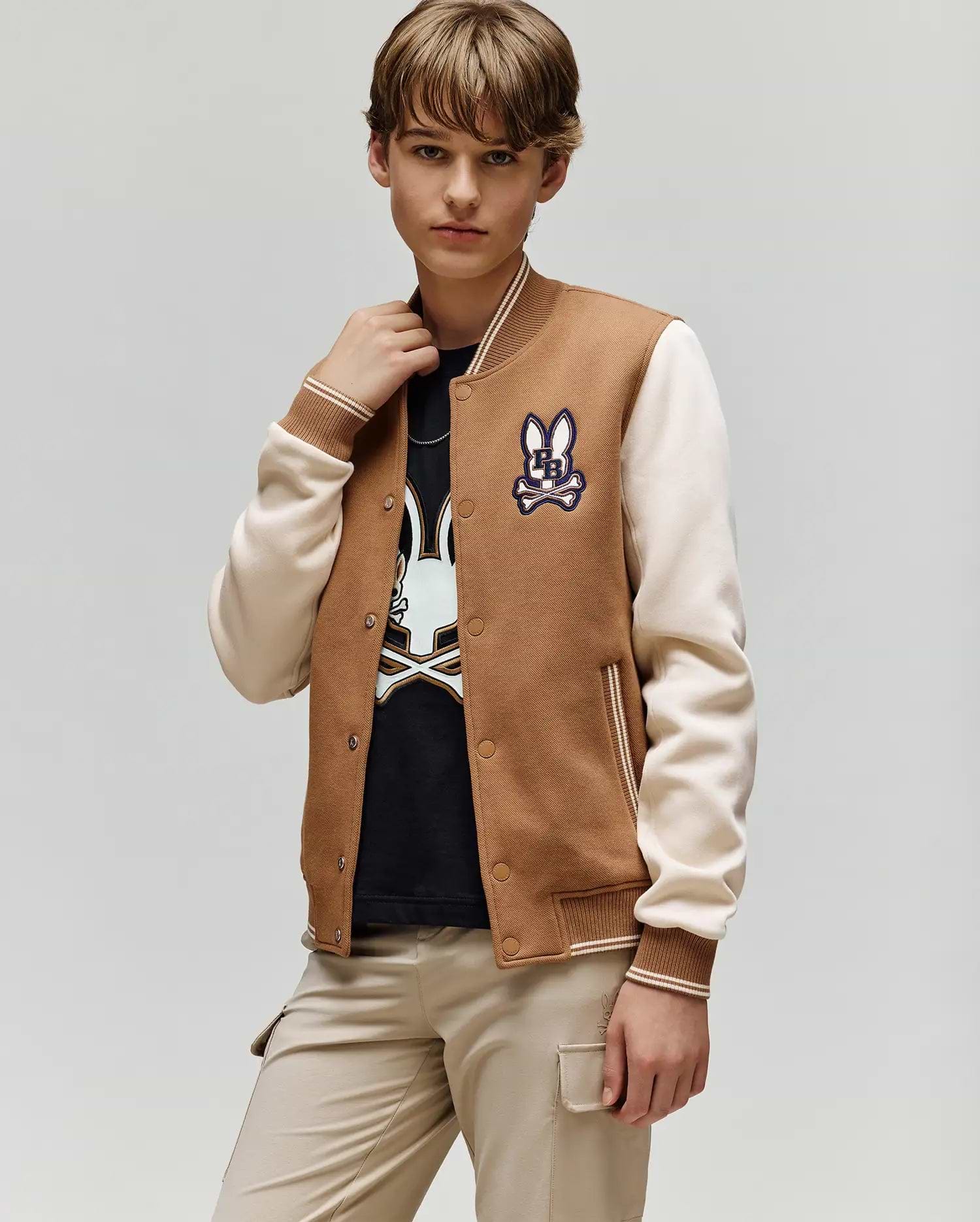 A young person wears the Psycho Bunny KIDS WADE KNIT BOMBER (KO1100001) in brown and beige, featuring a bunny patch, with a matching black bunny graphic shirt and beige pants, standing against a light gray background.