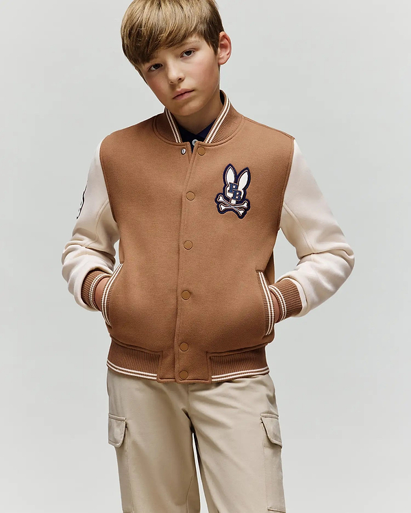 A young boy with light brown hair models the Psycho Bunny KIDS WADE KNIT BOMBER (KO1100001), a varsity-style jacket with a rabbit patch, paired with beige cargo pants, standing hands-in-pockets against a plain background.