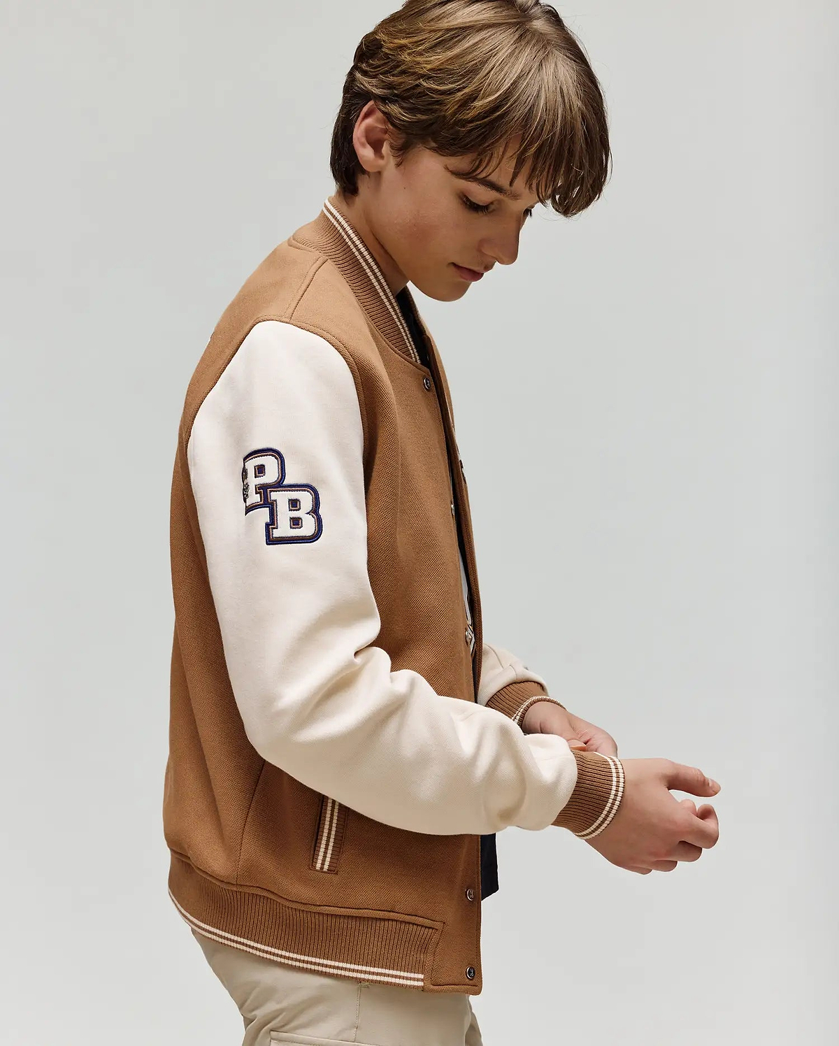 A young person with light brown hair looks down while adjusting their sleeve, wearing the Psycho Bunny KIDS WADE KNIT BOMBER (KO1100001), a varsity-style jacket with cream sleeves and "PB" lettering, set against a plain light background.