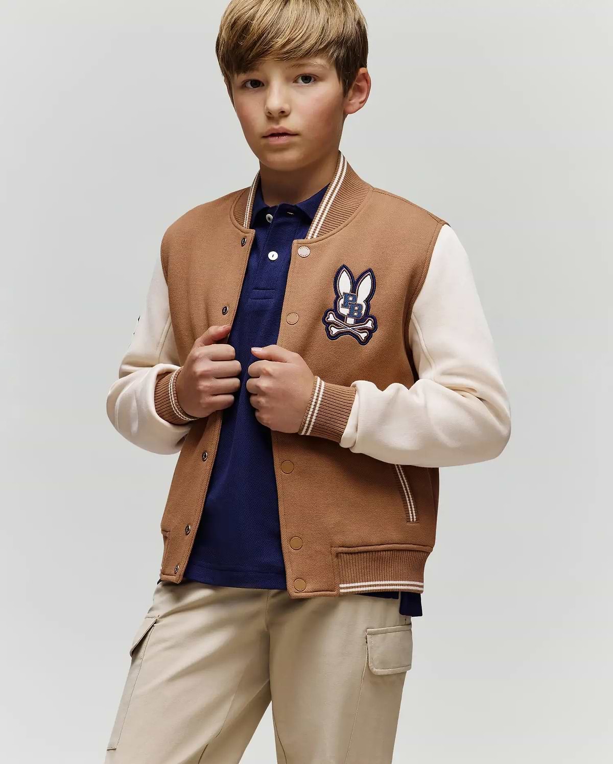 A young boy wears the Psycho Bunny KIDS WADE KNIT BOMBER (KO1100001) in tan and cream with a bunny patch, paired with a navy polo shirt and beige pants, standing against a plain background and looking at the camera neutrally.