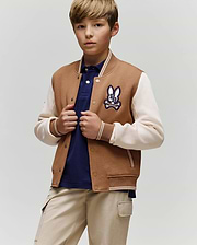 A young boy wears the Psycho Bunny KIDS WADE KNIT BOMBER (KO1100001) in tan and cream with a bunny patch, paired with a navy polo shirt and beige pants, standing against a plain background and looking at the camera neutrally.