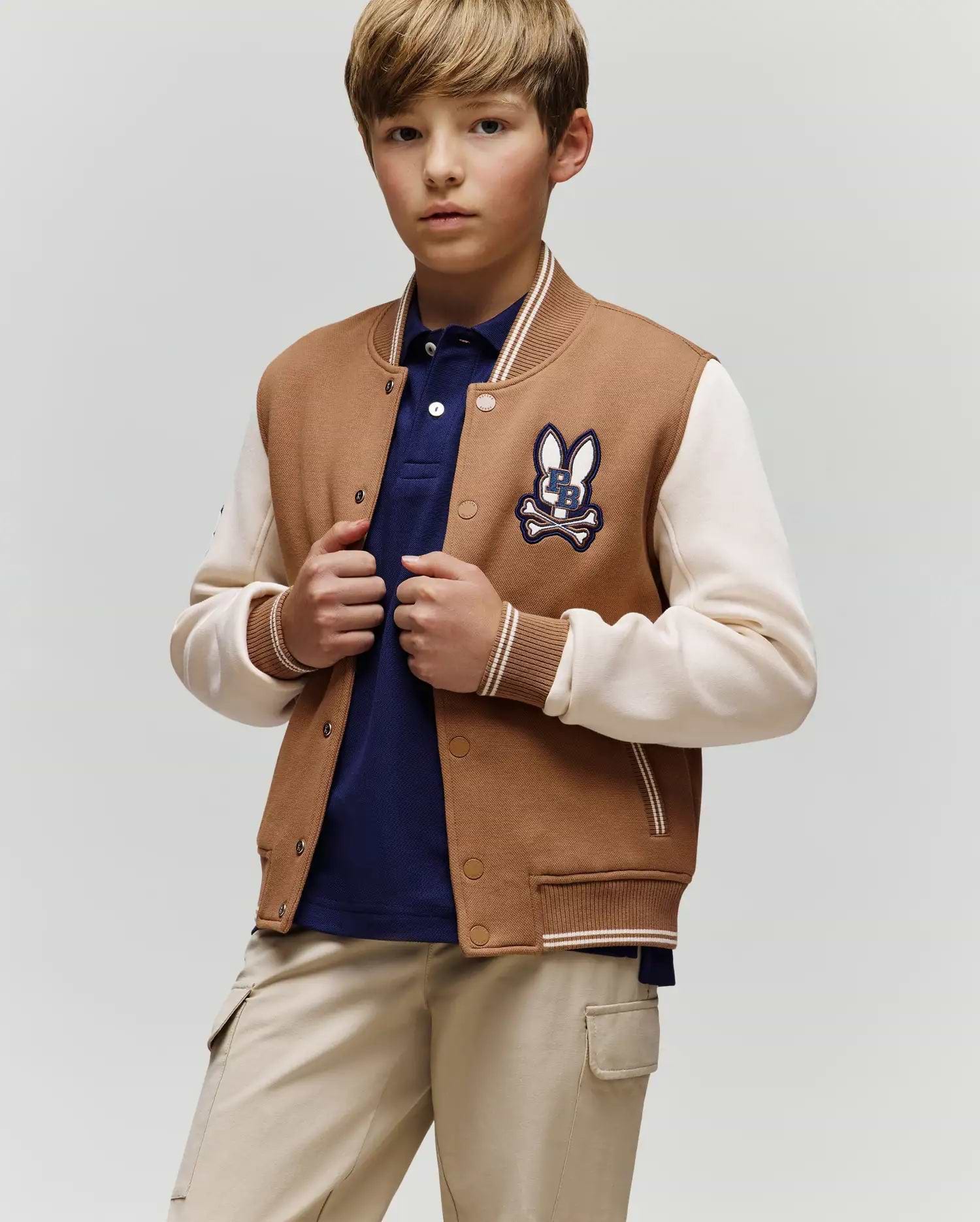 A young boy wears the Psycho Bunny KIDS WADE KNIT BOMBER (KO1100001) in tan and cream with a bunny patch, paired with a navy polo shirt and beige pants, standing against a plain background and looking at the camera neutrally.