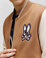 A child wears the Psycho Bunny KIDS WADE KNIT BOMBER (KO1100001) in tan and cream, featuring a bunny head and crossbones patch above "PB" on the chest. The child's face is partially out of frame.