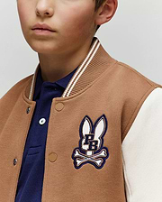 A young boy wears the Psycho Bunny KIDS WADE KNIT BOMBER (KO1100001) in tan and white, featuring a blue PB bunny and crossbones patch over a blue collared shirt. Only the lower half of his face is visible.