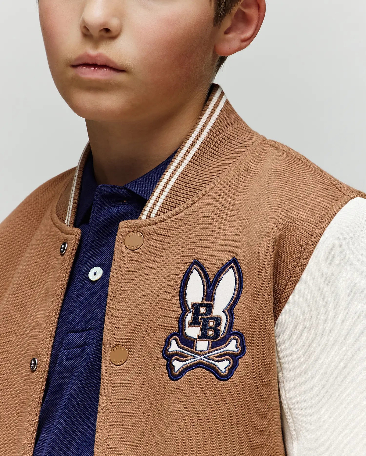 A young boy wears the Psycho Bunny KIDS WADE KNIT BOMBER (KO1100001) in tan and white, featuring a blue PB bunny and crossbones patch over a blue collared shirt. Only the lower half of his face is visible.