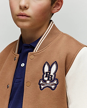 A young boy wears the Psycho Bunny KIDS WADE KNIT BOMBER (KO1100001) in tan and white, featuring a blue PB bunny and crossbones patch over a blue collared shirt. Only the lower half of his face is visible.