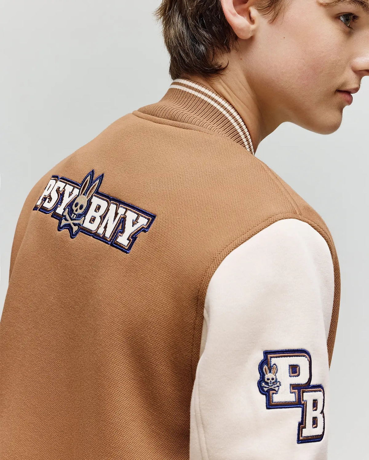 A young person models the Psycho Bunny KIDS WADE KNIT BOMBER (KO1100001), a premium brown and cream varsity jacket featuring "PSYBNY" and bunny logo on the back, plus a "PB" sleeve patch. Shown from the side, slightly turned away.