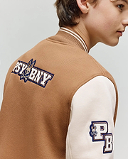 A young person models the Psycho Bunny KIDS WADE KNIT BOMBER (KO1100001), a premium brown and cream varsity jacket featuring "PSYBNY" and bunny logo on the back, plus a "PB" sleeve patch. Shown from the side, slightly turned away.