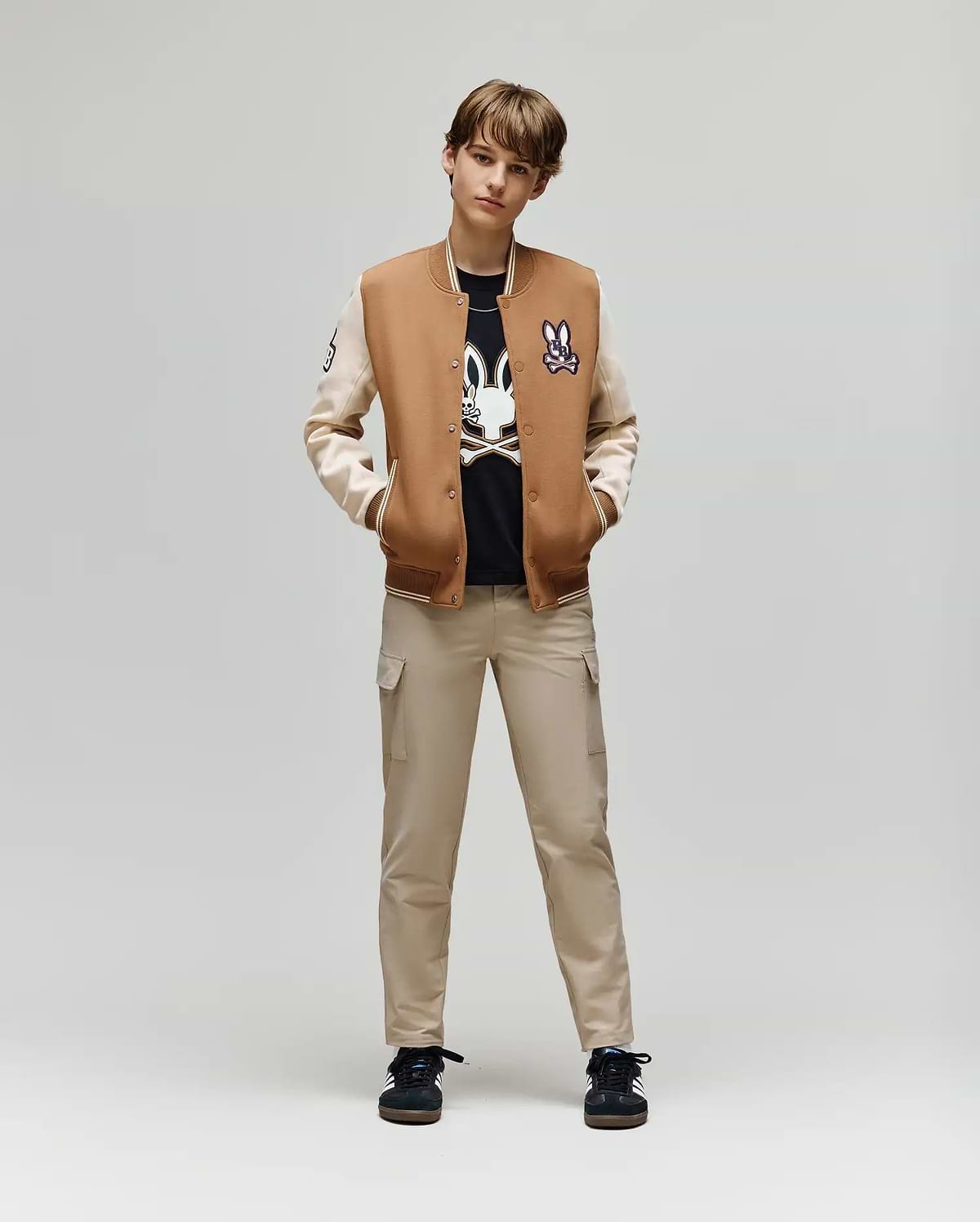 A young person stands facing forward, hands in pockets, wearing the Psycho Bunny KIDS WADE KNIT BOMBER (KO1100001) over a black graphic tee, beige cargo pants, and black sneakers against a plain light background.