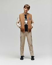 A young person stands facing forward, hands in pockets, wearing the Psycho Bunny KIDS WADE KNIT BOMBER (KO1100001) over a black graphic tee, beige cargo pants, and black sneakers against a plain light background.