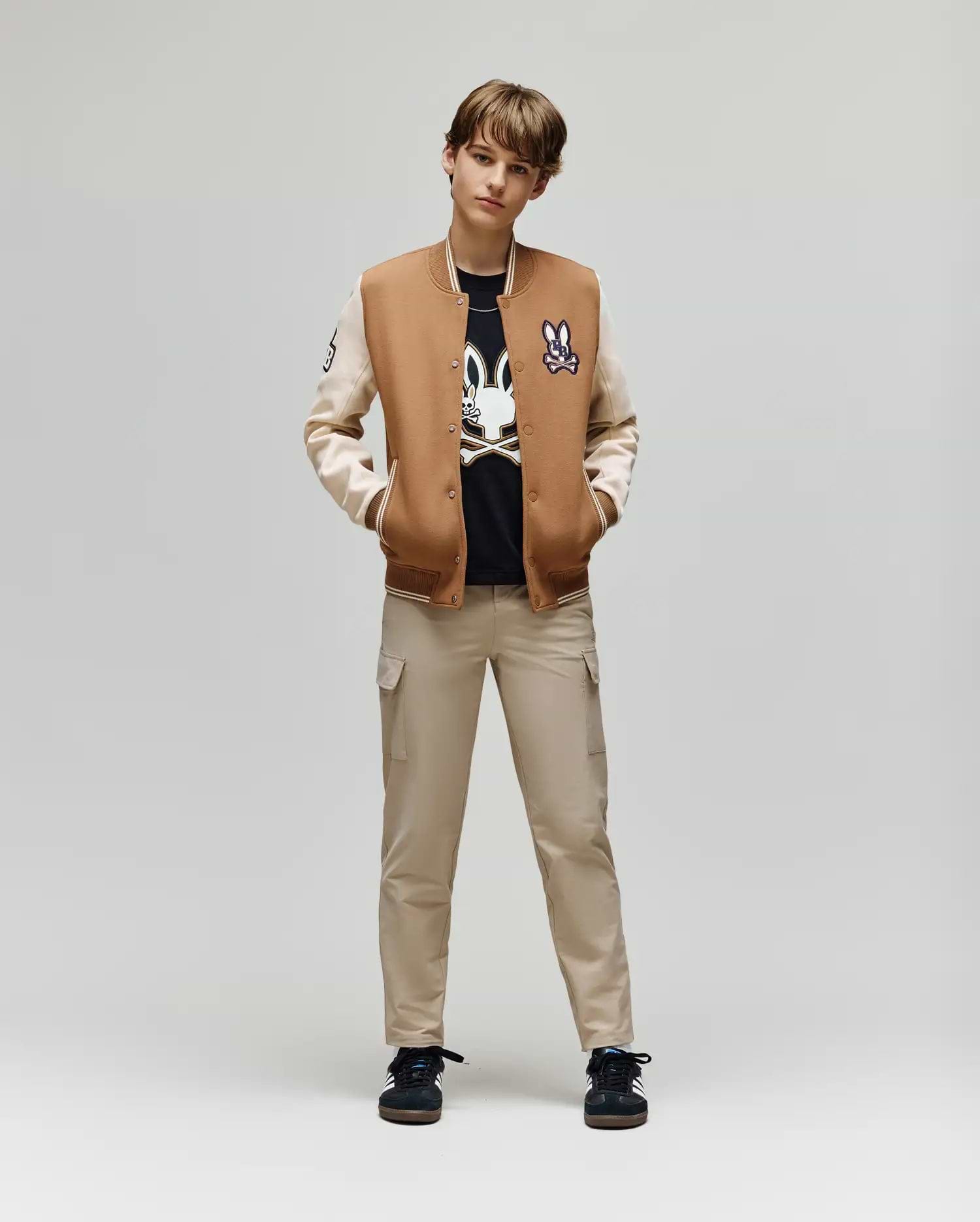 A young person stands facing forward, hands in pockets, wearing the Psycho Bunny KIDS WADE KNIT BOMBER (KO1100001) over a black graphic tee, beige cargo pants, and black sneakers against a plain light background.
