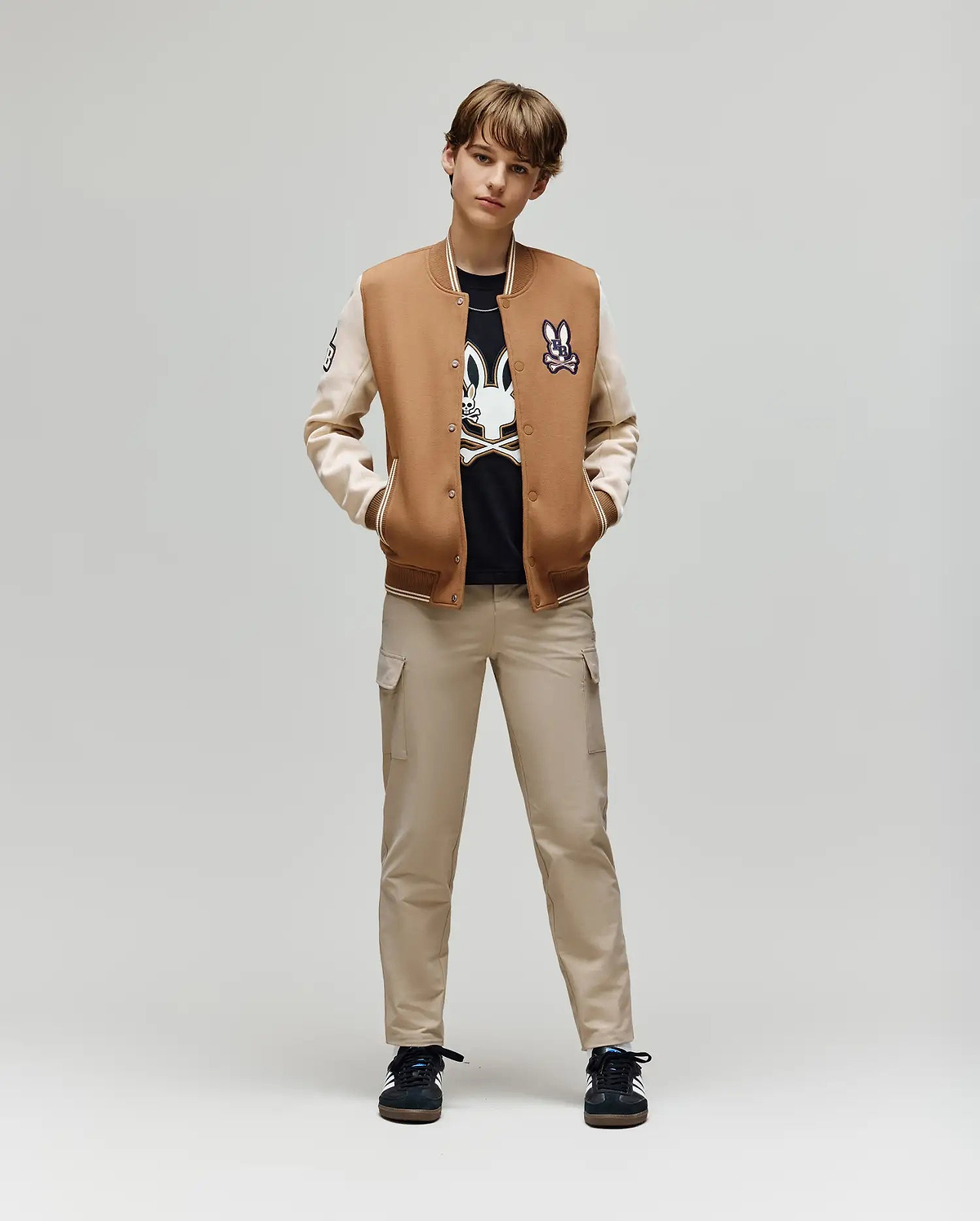 A young person stands facing forward, hands in pockets, wearing the Psycho Bunny KIDS WADE KNIT BOMBER (KO1100001) over a black graphic tee, beige cargo pants, and black sneakers against a plain light background.