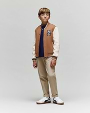 A young boy wears the Psycho Bunny KIDS WADE KNIT BOMBER (KO1100001), a premium cotton-blend tan and white varsity jacket with a cartoon patch, styled with a navy shirt, beige pants, and white sneakers. He stands calmly against a plain background.