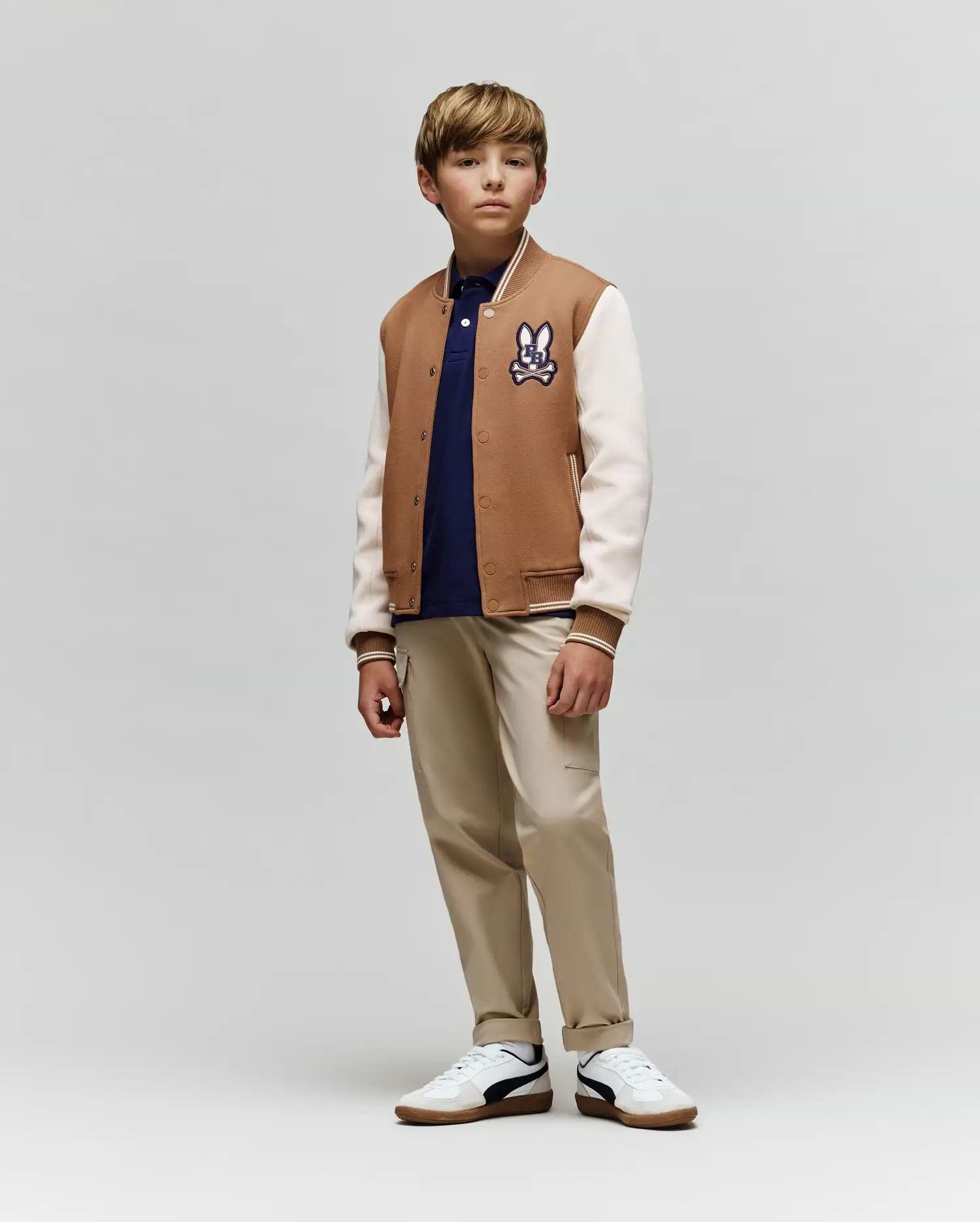 A young boy wears the Psycho Bunny KIDS WADE KNIT BOMBER (KO1100001), a premium cotton-blend tan and white varsity jacket with a cartoon patch, styled with a navy shirt, beige pants, and white sneakers. He stands calmly against a plain background.