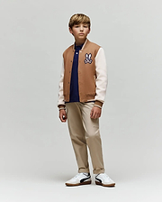 A young boy wears the Psycho Bunny KIDS WADE KNIT BOMBER (KO1100001), a premium cotton-blend tan and white varsity jacket with a cartoon patch, styled with a navy shirt, beige pants, and white sneakers. He stands calmly against a plain background.