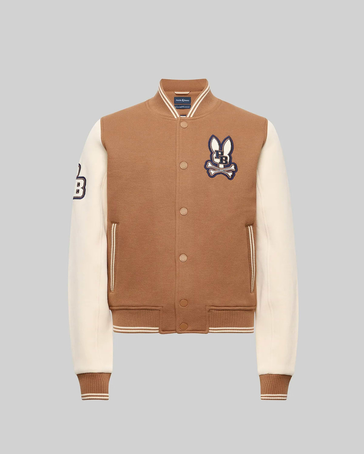 The Psycho Bunny KIDS WADE KNIT BOMBER (KO1100001) is a brown varsity jacket with cream sleeves, ribbed cuffs and hem, bunny patch on the chest, "B" patch on the sleeve, front pockets, and snap button closures. Made from a premium cotton blend.