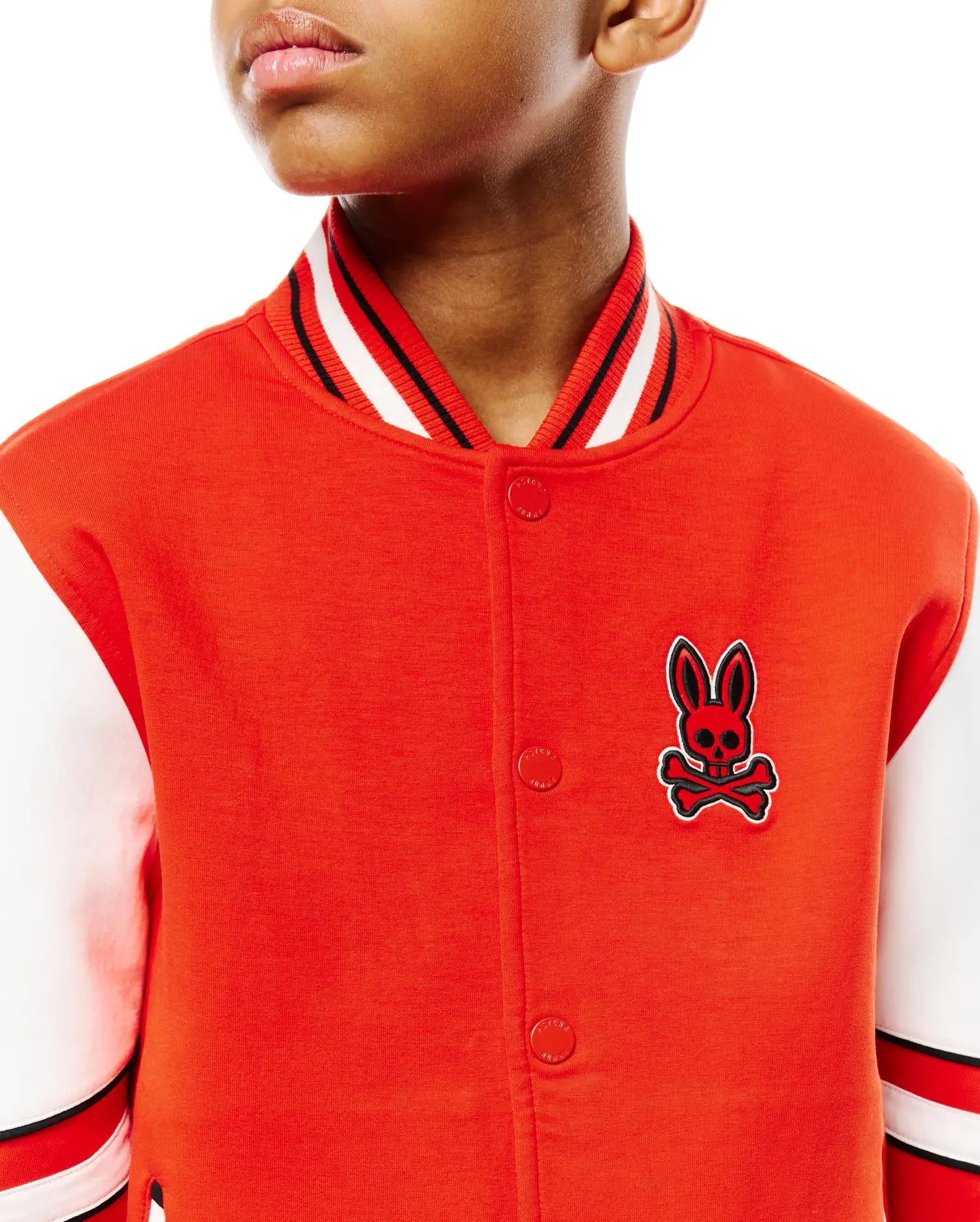 Kids Joey Knit Bomber, a vibrant orange jersey knit jacket with white sleeves, classic fit, featuring a large embroidered red and black Psycho Bunny skull logo on the left chest, and striped ribbed collar and cuffs.