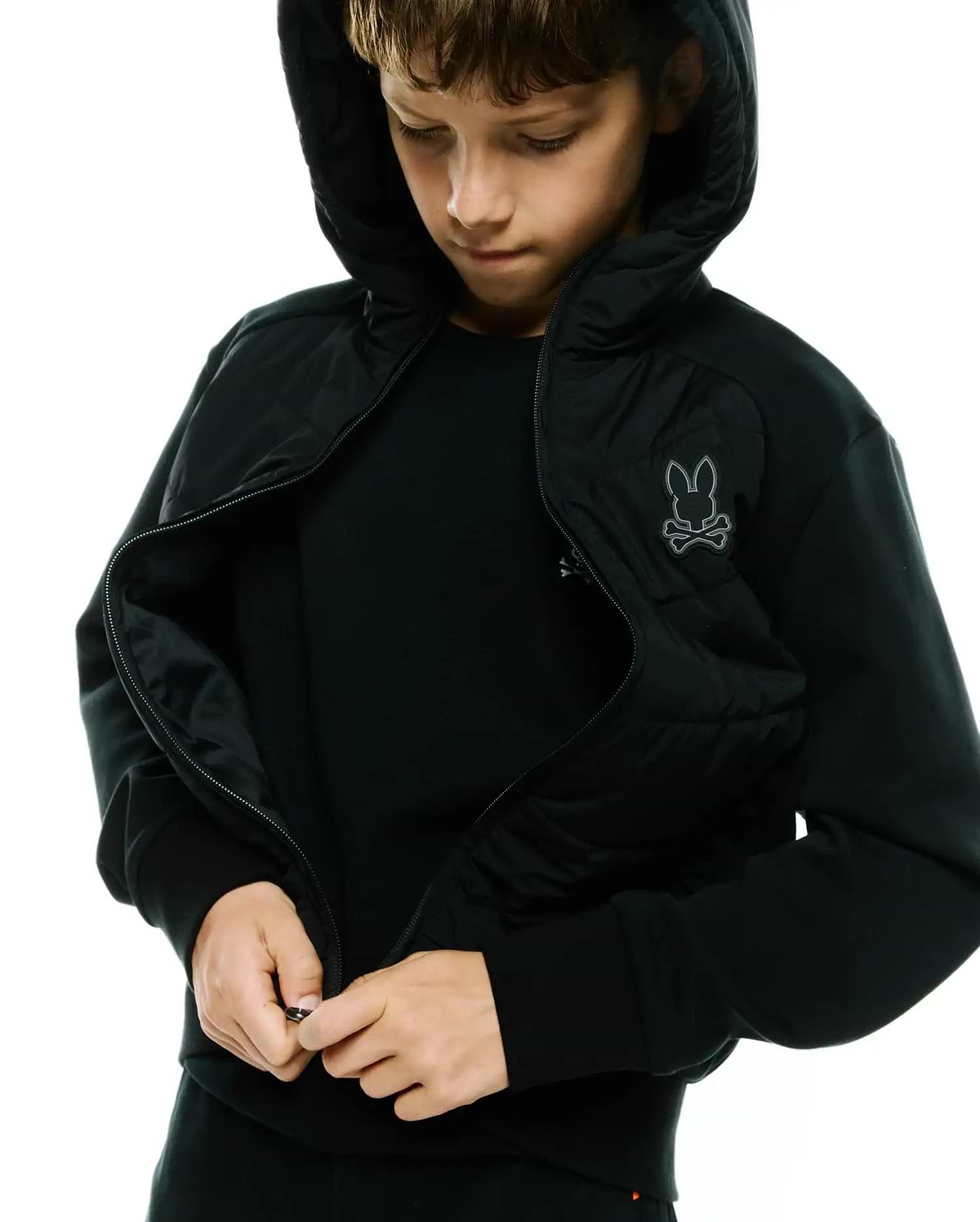 Kids Gordley Mix Media Jacket, a black hooded full-zip jacket with a quilted front panel, soft jersey sleeves, and an embroidered grey Psycho Bunny skull logo on the left chest.
